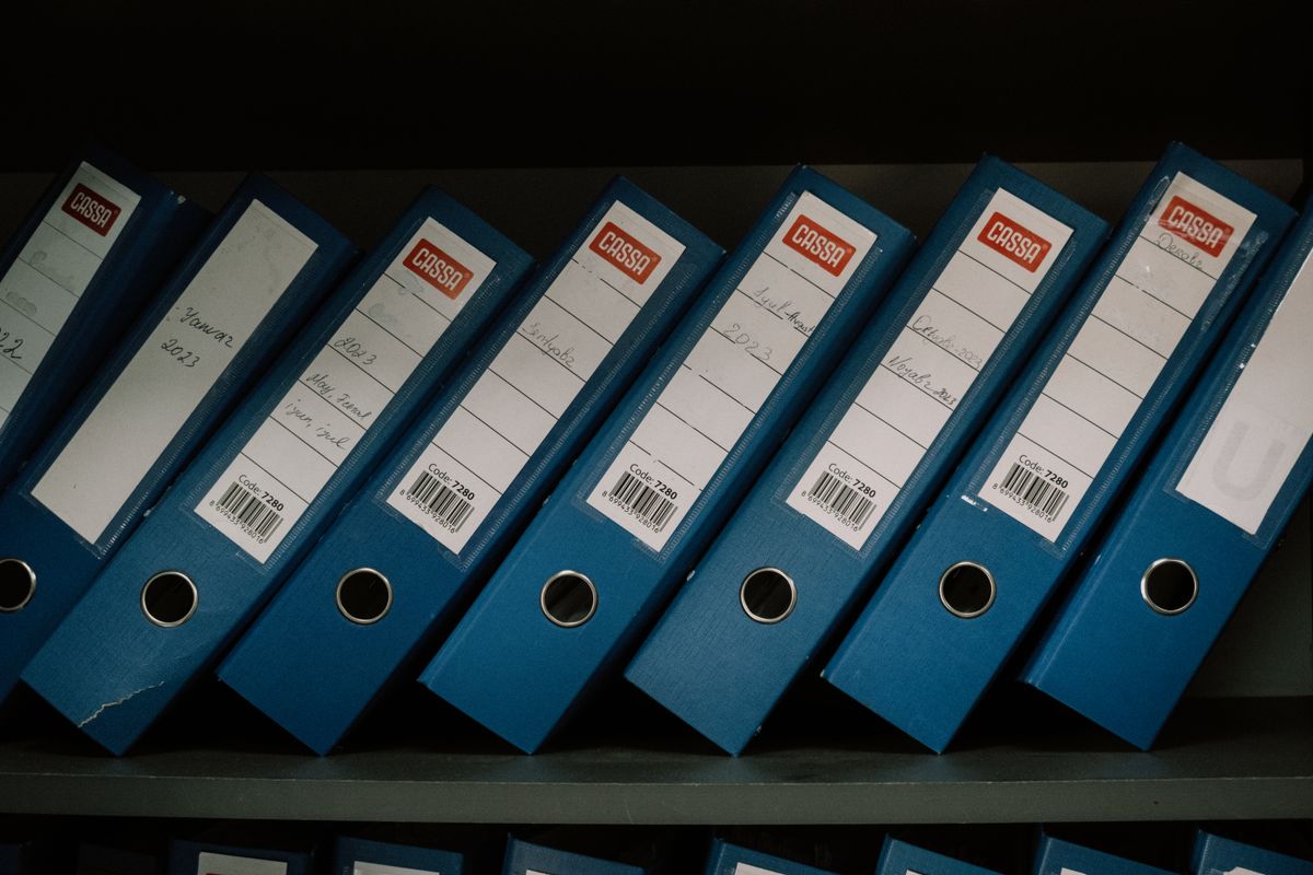 Row of labelled and dated archive binders on a shelf, the paper records a document management system replaces